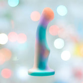 The Avant Opal Dreams Dildo - Aqua by Avant is a pastel-colored silicone sculpture with a smooth, curved shape and flat base, showcased against a softly blurred background with gentle bokeh light effects.